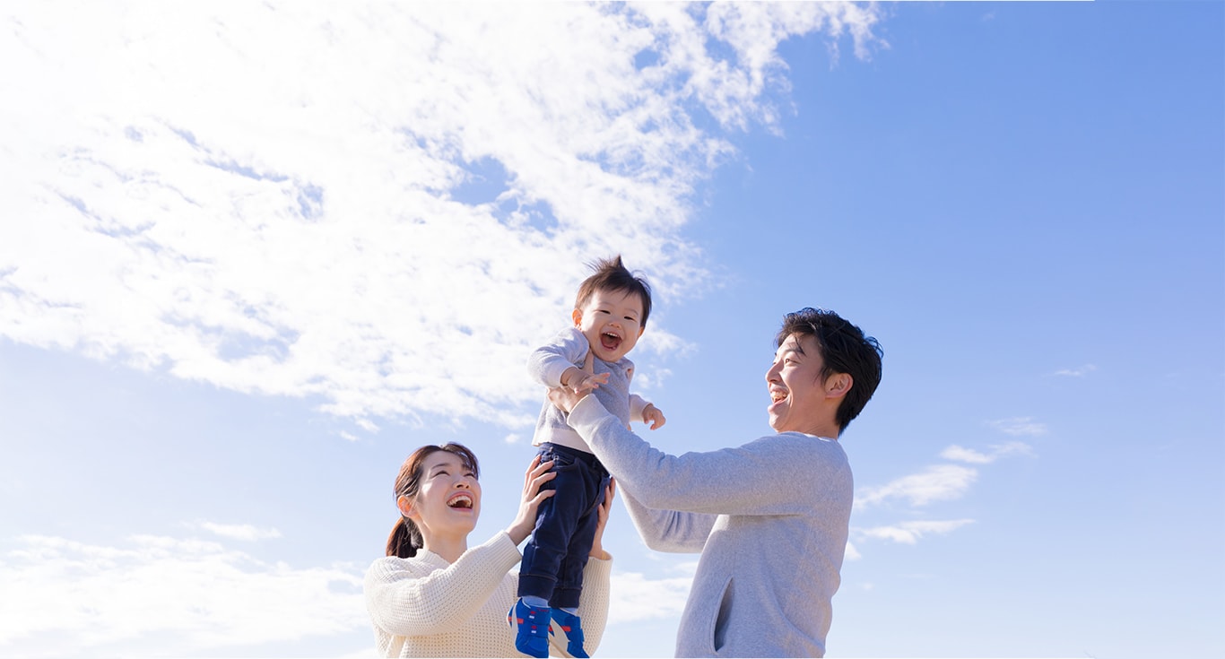 親子三人の家族写真。父親が子供を抱き上げている。背景には青空。